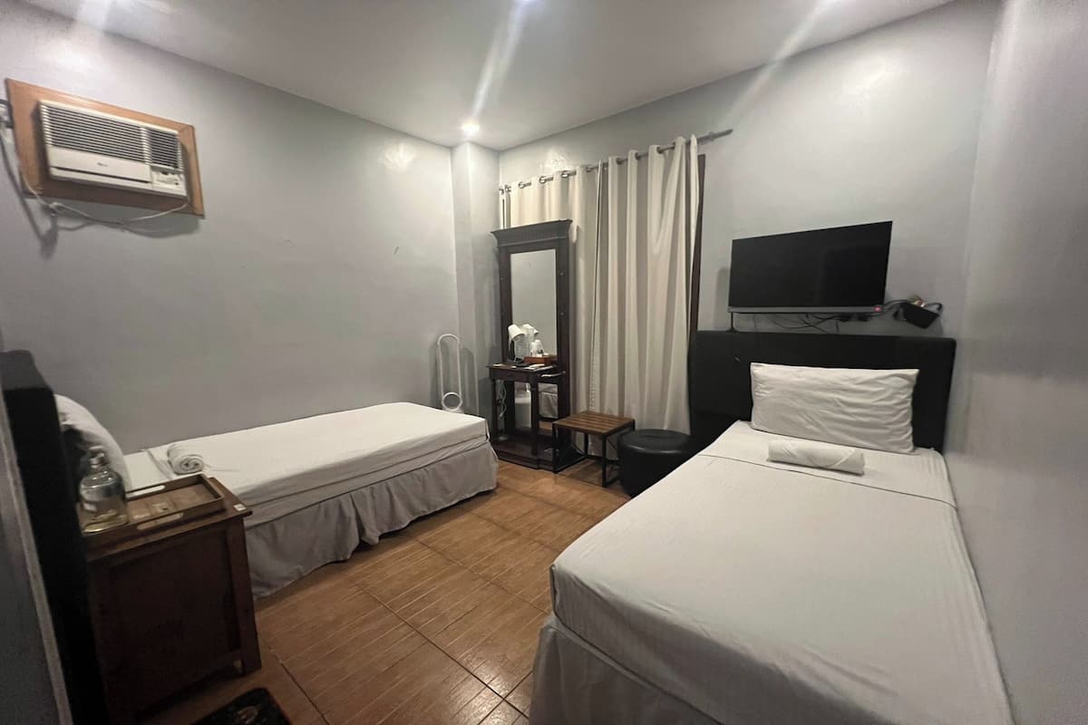 A hotel room with two single beds, a TV, a mirror, a small table, and an air conditioning unit.
