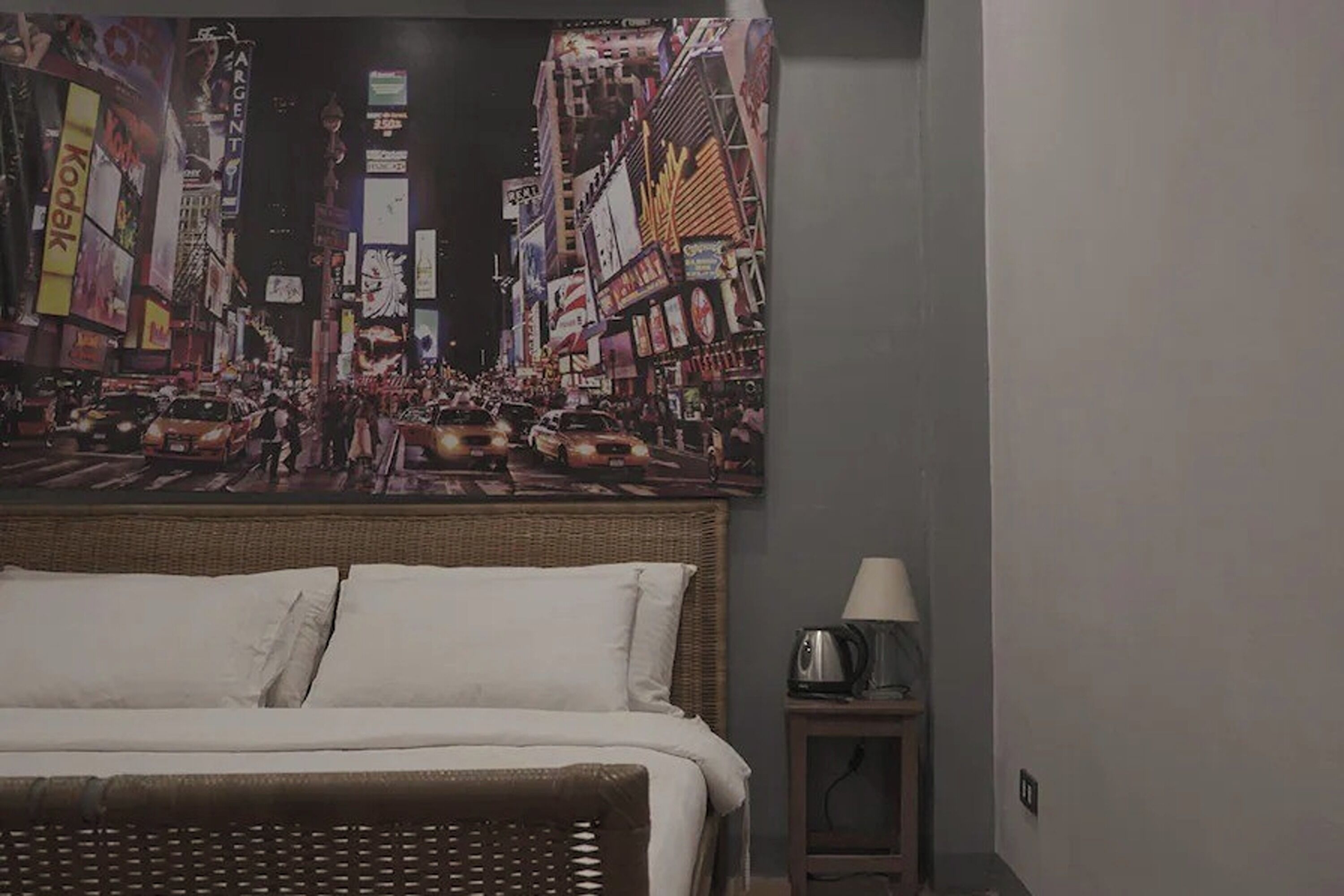 A hotel room with a large cityscape painting on the wall, a bed with white linens, and a small bedside table with a lamp.