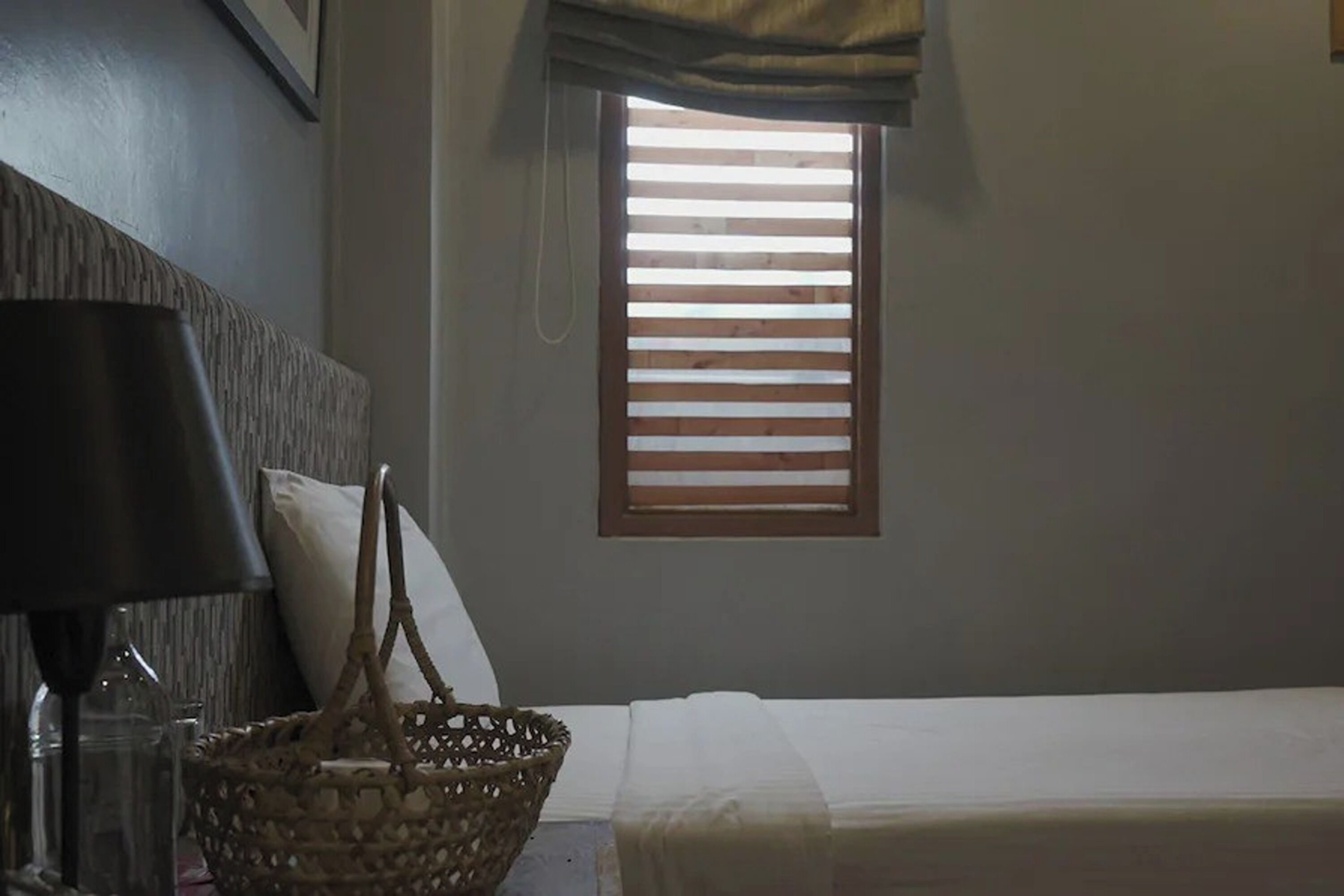 Un dormitorio con cama, lámpara de noche, cesta tejida y ventana con persianas de madera.