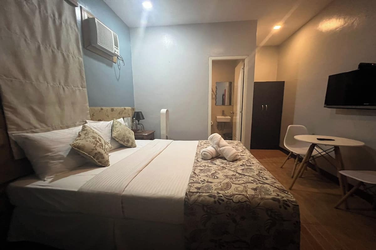 A hotel room with a bed, a desk, a chair, a TV, and an air conditioning unit.