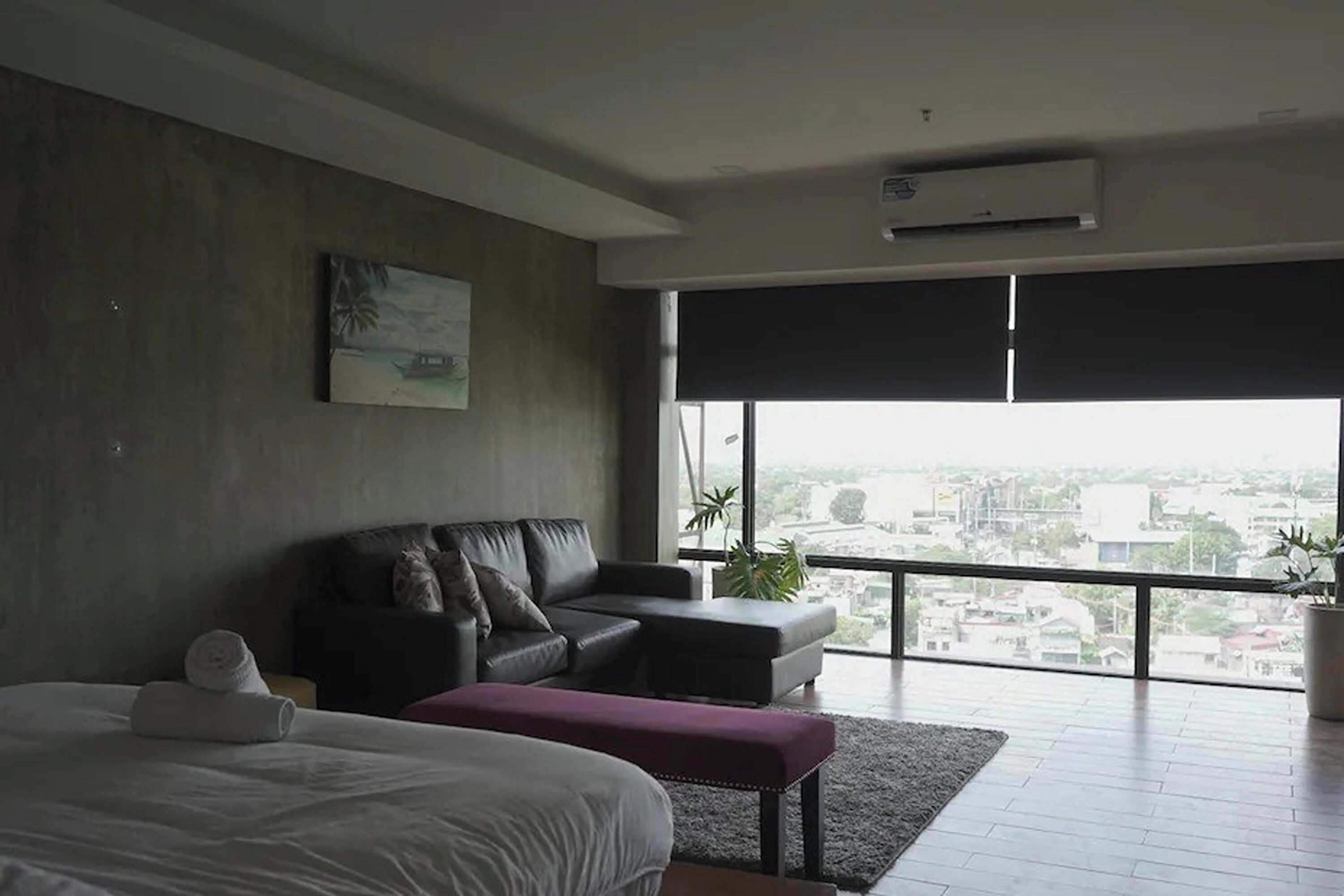A modern hotel room with a large bed, a sofa, a bench, and a city view through large windows.