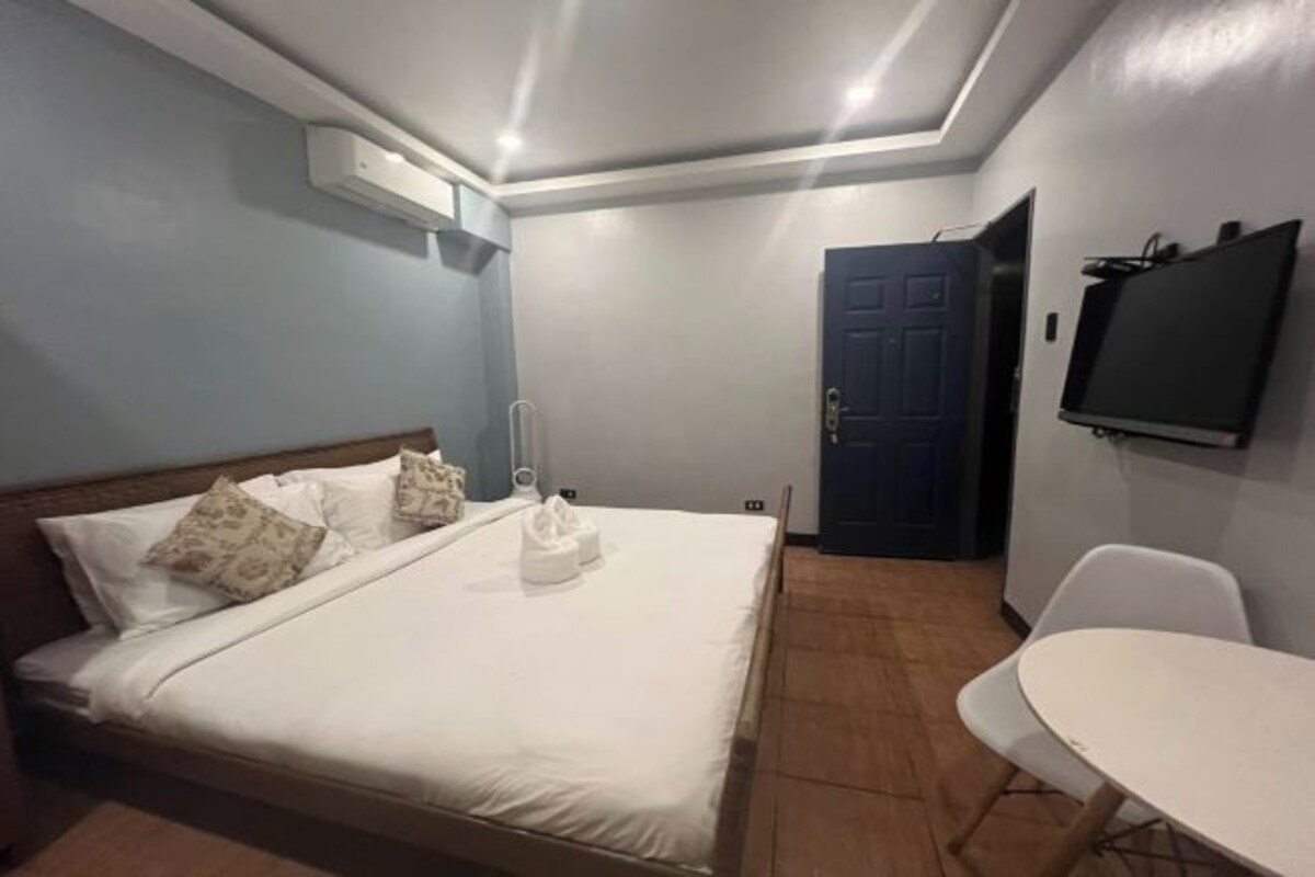 Un dormitorio con una cama, un televisor montado en la pared, una puerta y una mesita.