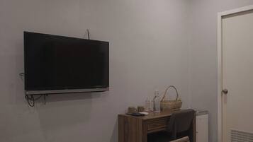 A room with a flat-screen TV mounted on the wall, a wooden desk with a chair, a small refrigerator, and a basket on the desk.