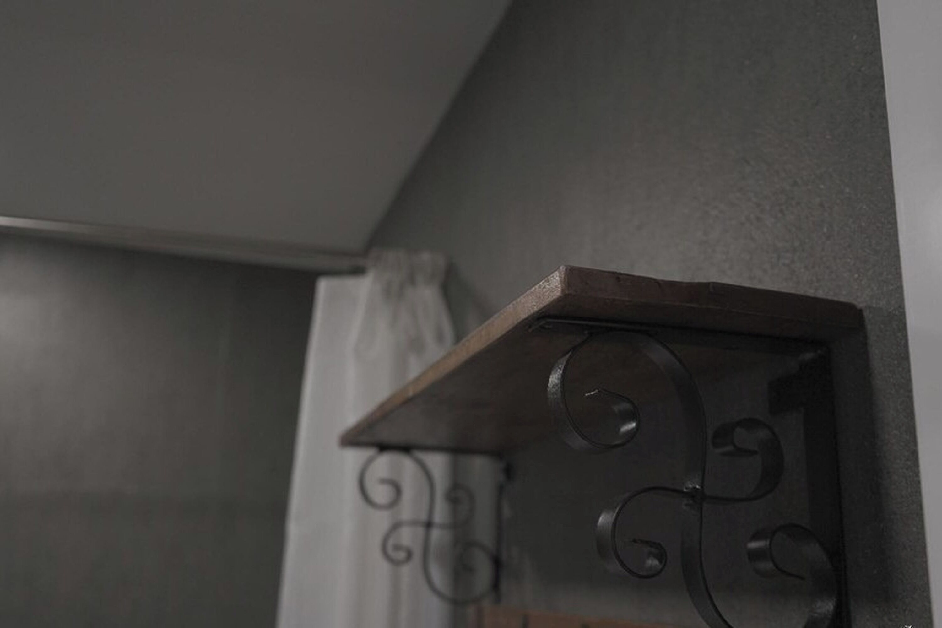 Rincón de una habitación con un estante de madera oscura contra una pared gris, un adorno metálico y una cortina parcialmente visible.