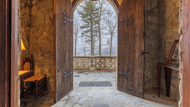A stone-walled room with a wooden bench, a small table, and a view of trees through an arched doorway.