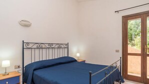 A bedroom with a metal bed frame, a blue bedspread, a bedside table with a lamp, and a view through a glass door to an outdoor area. - Li Cossi by Interhome (Costa Paradiso)