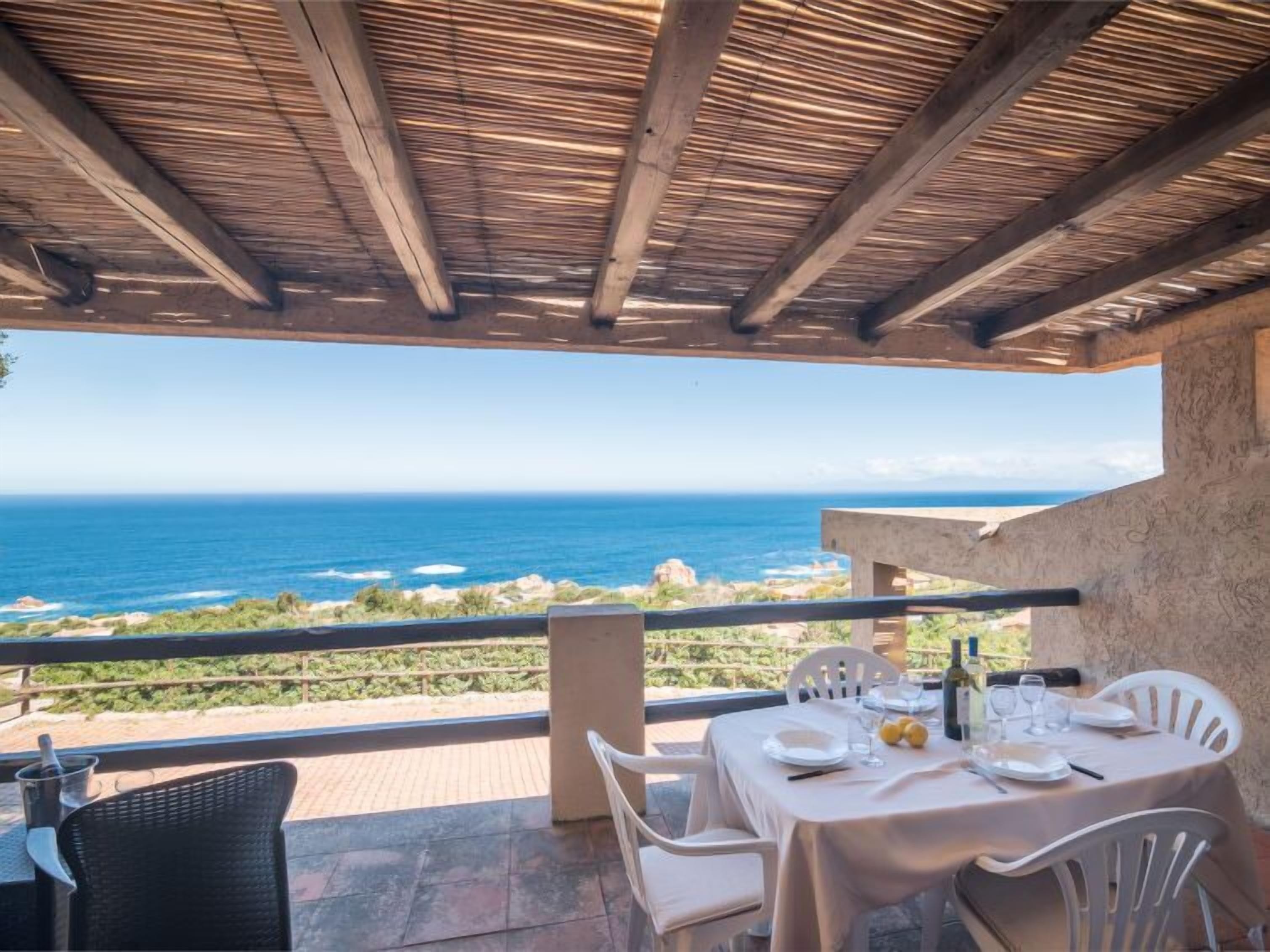 Ein Essbereich im Freien mit Blick auf das Meer, mit einem für zwei Personen gedeckten Tisch, weißen Stühlen, einer Pergola darüber und einer Steinmauer.