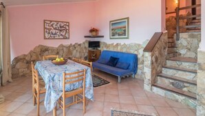 A cozy living room with a stone wall, a blue sofa, a dining table with chairs, and a staircase. - Li Cossi by Interhome (Costa Paradiso)