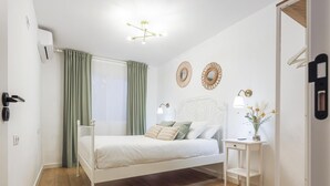 Una camera da letto con un letto bianco, tende verdi, un comodino con una lampada e uno specchio decorativo al muro.