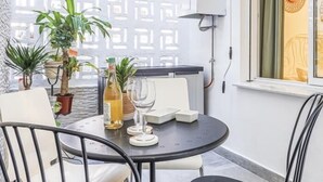 Un balcone con un tavolo rotondo, sedie, piante in vaso, un termosifone e una finestra con un oggetto decorativo.