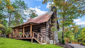 A rustic log cabin with a wooden porch and stone chimney, surrounded by trees and a grassy area. - Retreat that will have you wanting more RELAxATION (Sevierville)