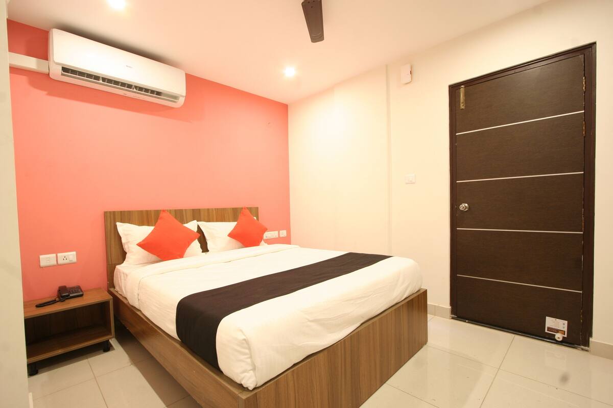 A hotel room with a bed, a nightstand, an air conditioning unit, and a door.