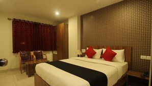 A hotel room with a large bed, a wooden headboard, a nightstand, a desk with a chair, and a window with curtains. - PPH Living Nagavara Lake Sai Inn (Bengaluru)