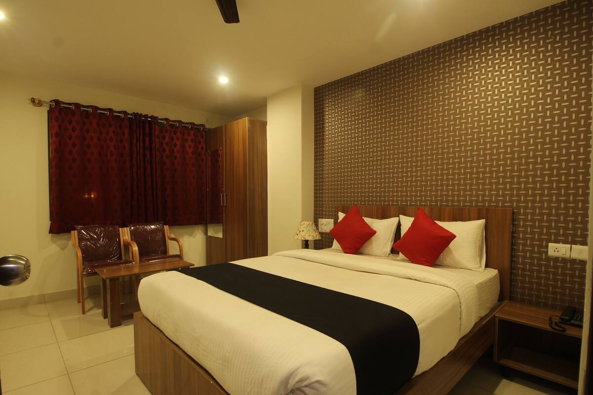 A hotel room with a bed, a wooden headboard, a nightstand, a desk with a chair, a mirror, and a window with curtains.