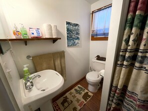 Un baño con lavamanos, inodoro y cortina de ducha. Hay artículos de aseo en un estante y una toalla colgada en la pared.