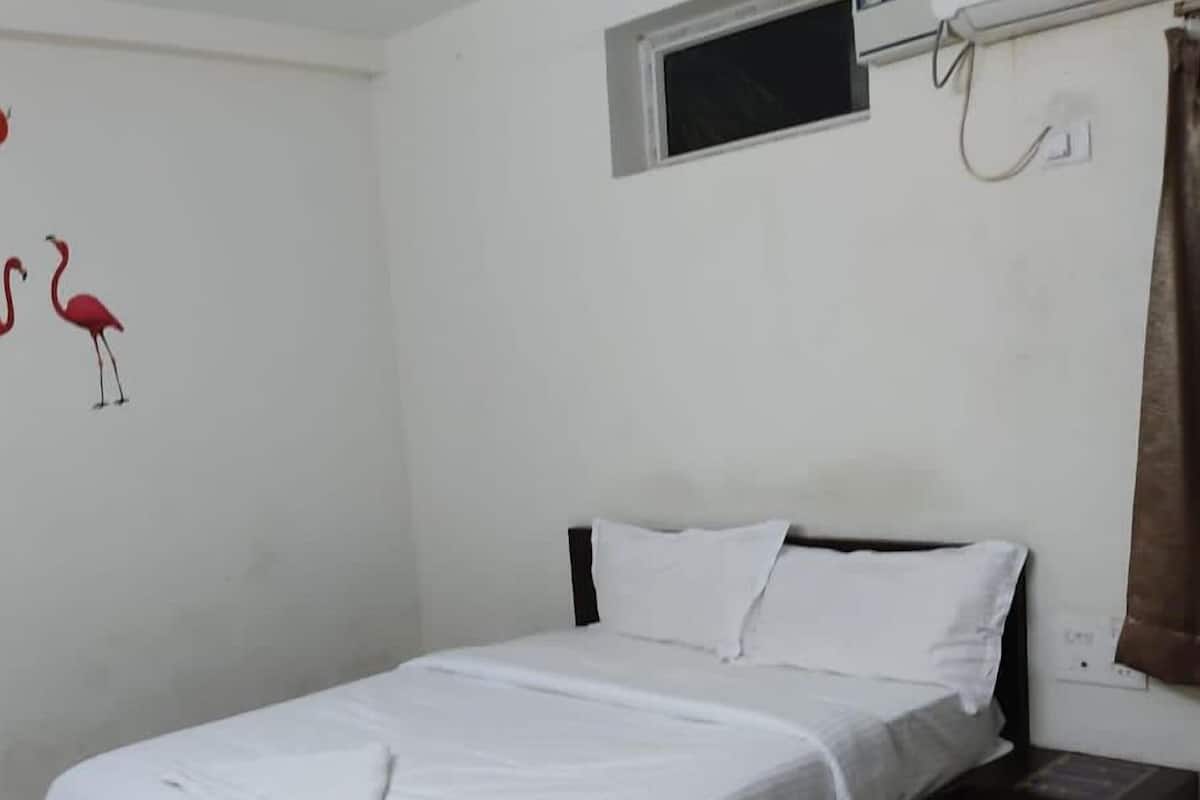 A single bed with white bedding in a room with white walls, a small window, and a wall-mounted light fixture.