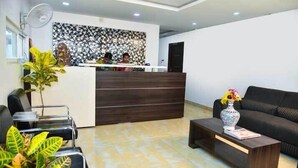 A reception area with a dark wooden reception desk, a black sofa, and a coffee table with a vase. - PPH Living Bhimas (Tada)