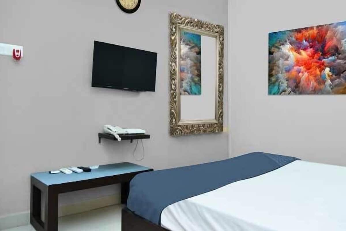 A hotel room with a bed, a TV, a bedside table with a phone, a mirror, and a painting on the wall.