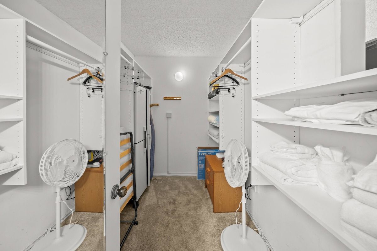 Ein begehbarer Kleiderschrank mit weißen Regalen, einem Ventilator und einer Kiste Handtücher der Marke Lasko.