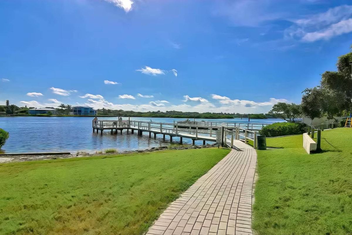 Uma propriedade à beira-mar com um calçadão pavimentado que leva a um cais, área gramada e vista desimpedida para a água.