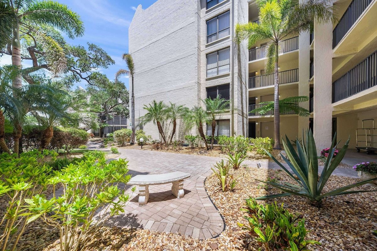 A courtyard with a central seating area, surrounded by palm trees and various plants, leading to a building with multiple balconies.