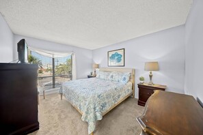 A bedroom with a bed, a dresser, a nightstand, a lamp, and a window with blinds.