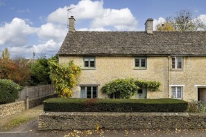 A stone house with a steeply pitched roof, multiple chimneys, and a small front garden with a hedge and a tree. - Family friendly holiday cottage in the Cotswolds with a superb garden - Winterberry Cottage (Barnsley, near Cirencester)