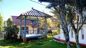 A covered outdoor seating area with a swing set, surrounded by a well-maintained lawn and trees. - Classic Herman by Broga (Cipanas)