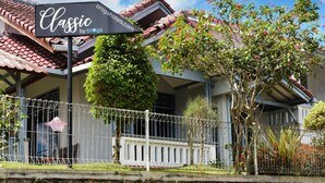 A building with a sign reading "Classic by brioga hotels.com" and a white fence with greenery. - Classic Herman by Broga (Cipanas)