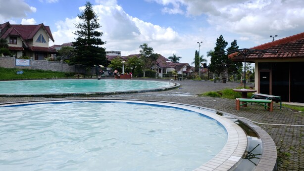 A swimming pool with a tiled deck, surrounded by a park area with benches and trees. - Classic Herman by Broga (Cipanas)