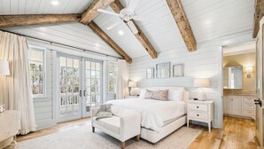 A bedroom with a large bed, a nightstand, a chair, a ceiling fan, and a view of the outdoors through a sliding door.