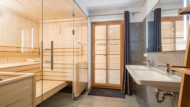 Moderni sauna, jossa on puuseinät ja lasiovi, suuri ikkuna, jossa on sälekaihtimet, sekä erillinen kylpyhuone, jossa on pesuallas ja peili.