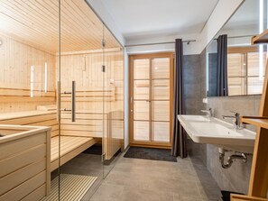 A modern sauna with wooden walls and a glass door, a large window with blinds, and a separate bathroom with a sink and mirror.