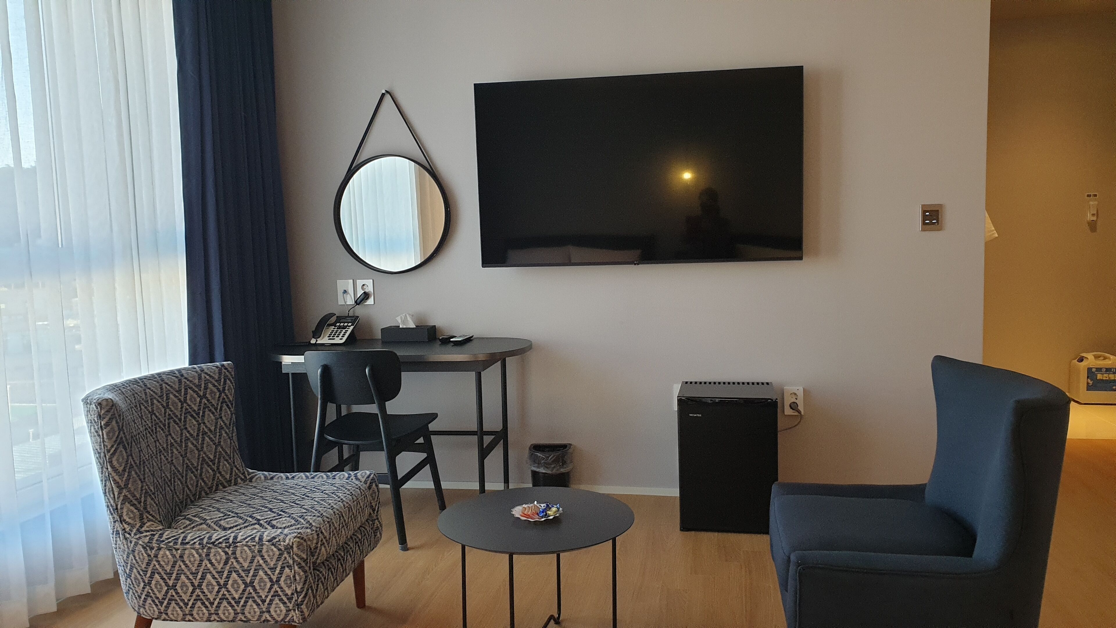 A compact living space with a patterned armchair, a dark armchair, a small round table, a desk with a computer and phone, a TV mounted on the wall, and a compact refrigerator.