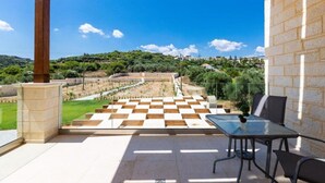 Un porxo cobert amb un paviment de pissos quadriculats, una taula i cadires, amb vista a un jardí ben cuidat i a les colines allunyades.