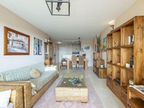 Een gezellige woonkamer met een bank, een salontafel en een boekenkast. Er is een eethoek met stoelen en een tafel, en een keuken op de achtergrond.