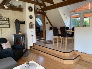 Un salon chaleureux avec une cheminée, un coin repas avec des chaises en bois et une cuisine visible à travers une porte ouverte.