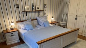 Ein Schlafzimmer mit einem hölzernen Bettgestell, weißer Bettwäsche, zwei Nachttischen mit Lampen, einem zusätzlichen Tisch und einem Wandregal mit Bilderrahmen.