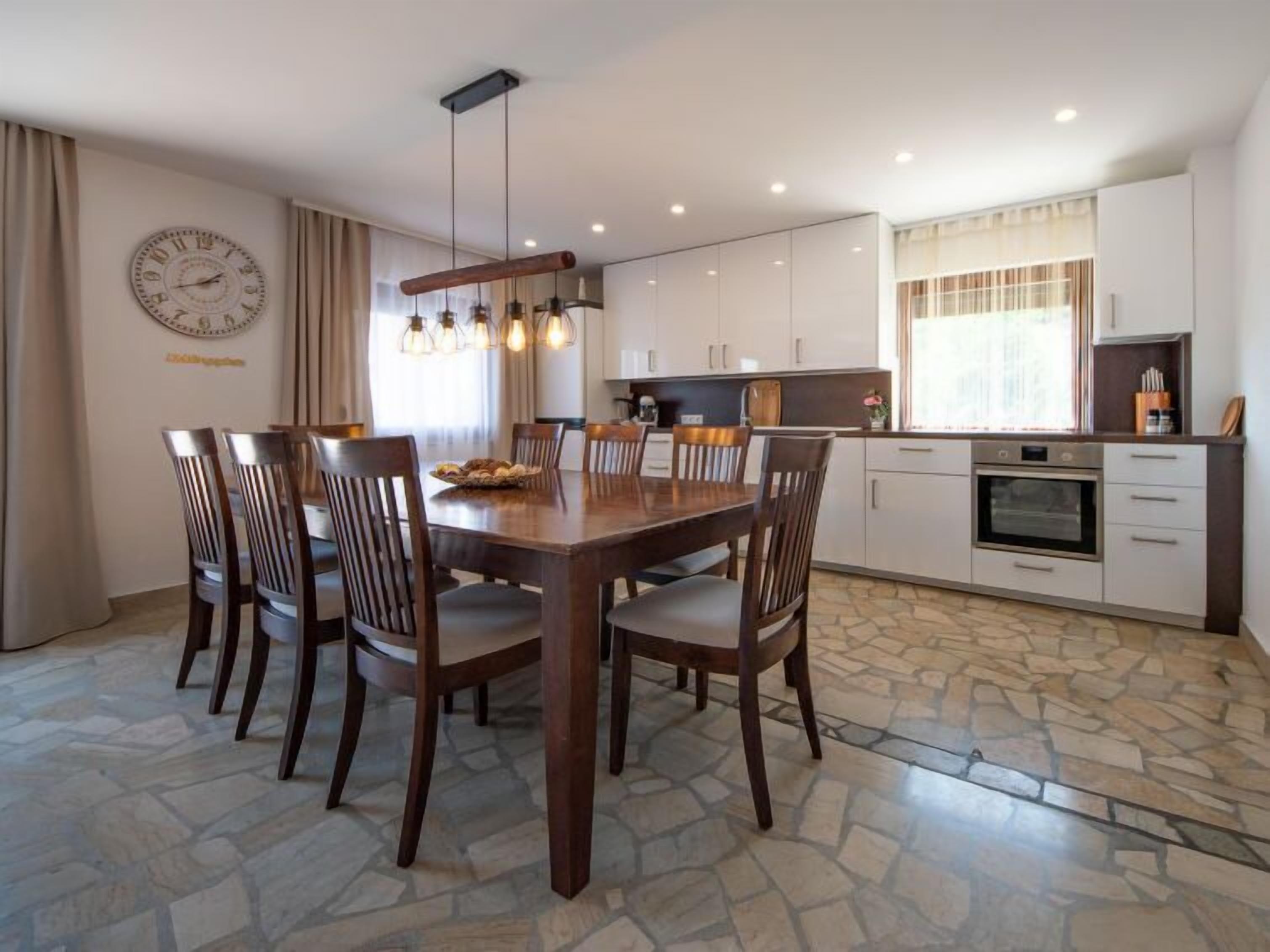 A modern kitchen with a dining area, featuring a large wooden table, stone flooring, and pendant lighting.