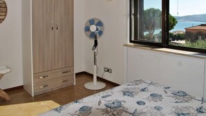 Una camera da letto con un letto, una ventola da pavimento, un armadio, una finestra vista mare e una lampada a parete.