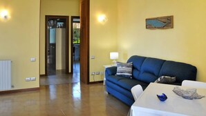 Un soggiorno con un divano blu, un tavolo bianco e una lampada. Si apre una porta verso un'altra stanza.