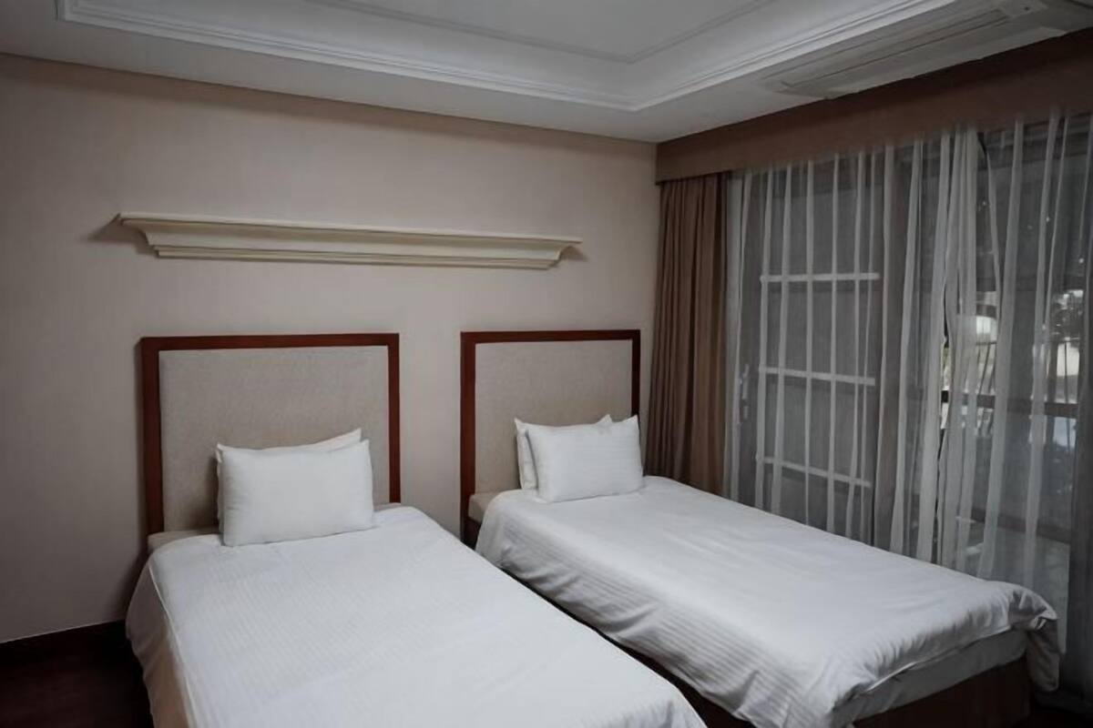 A hotel room with two single beds, a large window with sheer curtains, and a ceiling light fixture.