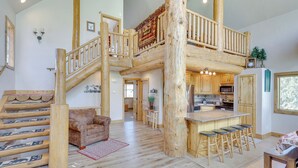A spacious living area with a wooden staircase, a cozy leather sofa, and a kitchen with wooden bar stools. - Rustic Sterling Cabin: Walk to Palisade Lake! (Sterling)