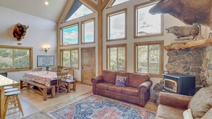 A spacious living room with a large window, a stone fireplace, and a mounted deer head. - Rustic Sterling Cabin: Walk to Palisade Lake! (Sterling)