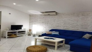 A living room with a blue sofa, a TV on a white stand, a small table, and a woven pouf. - Apartment Barbara (Vir)