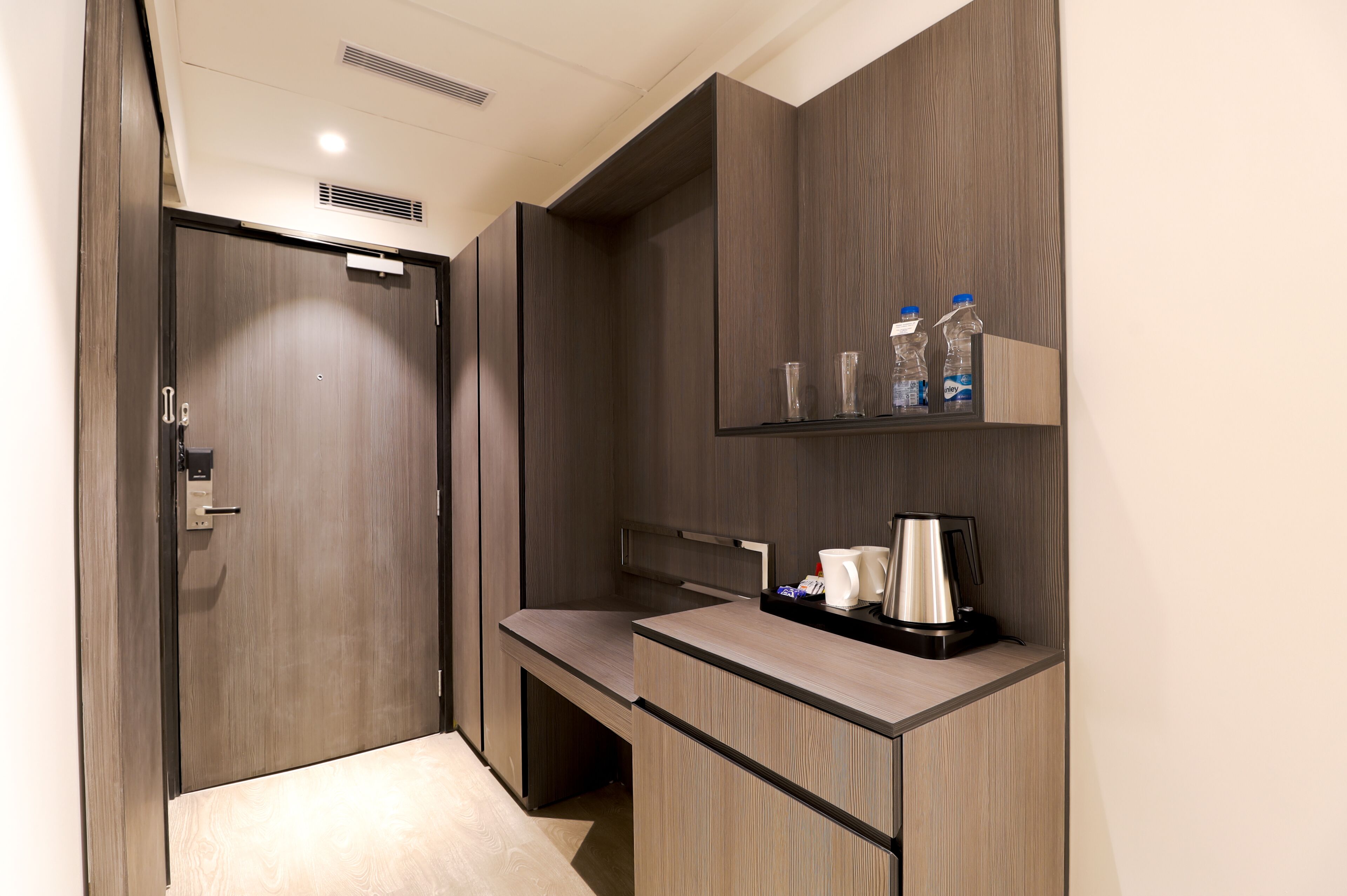A compact hotel room with a wooden wardrobe, a small desk with a coffee maker, and a shelf with bottles and glasses.