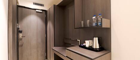 A compact hotel room with a wooden wardrobe, a small desk with a coffee maker, and a shelf with bottles and glasses.