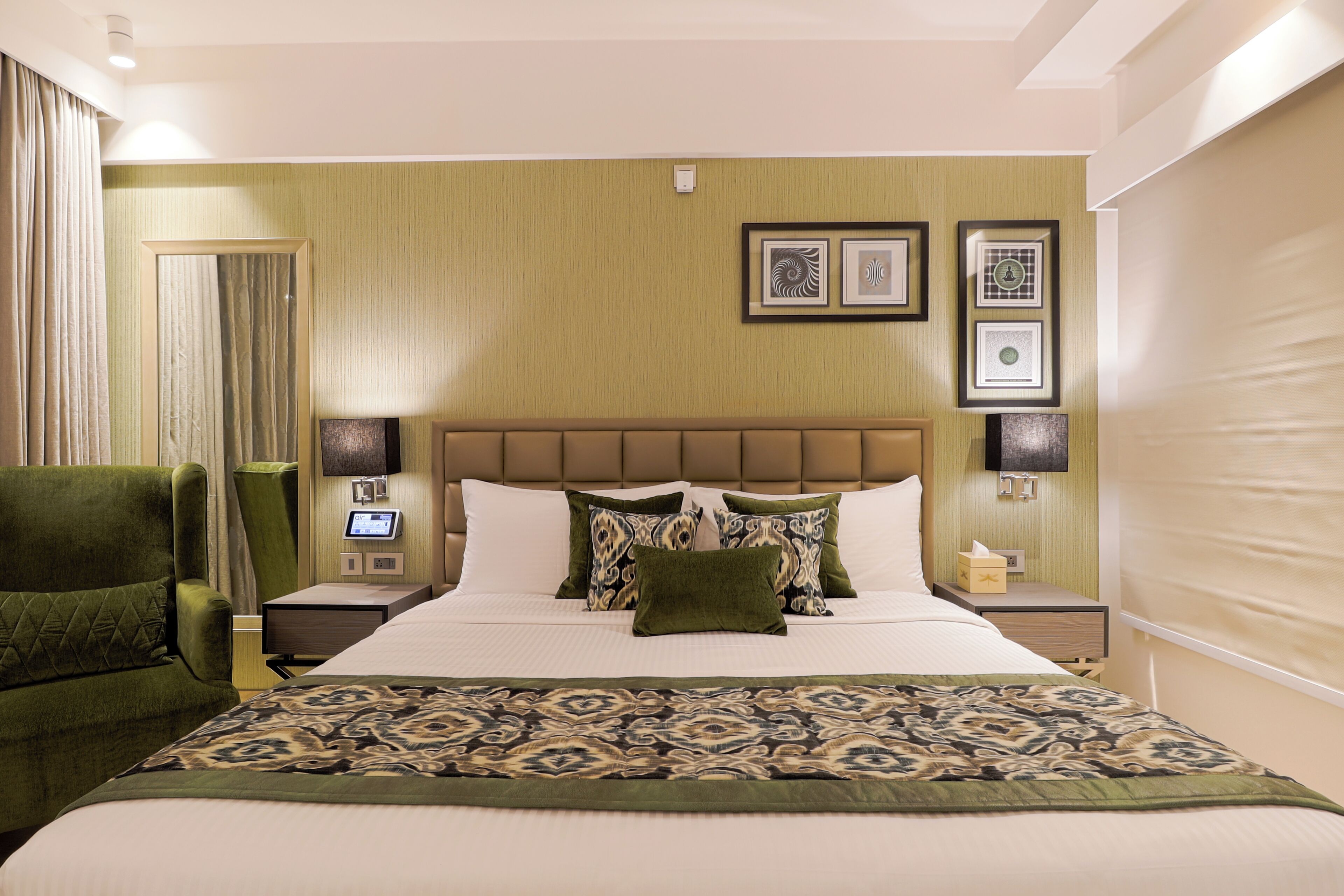 Une chambre d’hôtel avec un grand lit, un fauteuil vert, une table de chevet avec une lampe et des œuvres d’art encadrées accrochées au mur.