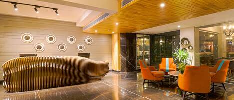 A modern lobby with a wooden reception desk, a seating area with orange chairs, and a polished marble floor.