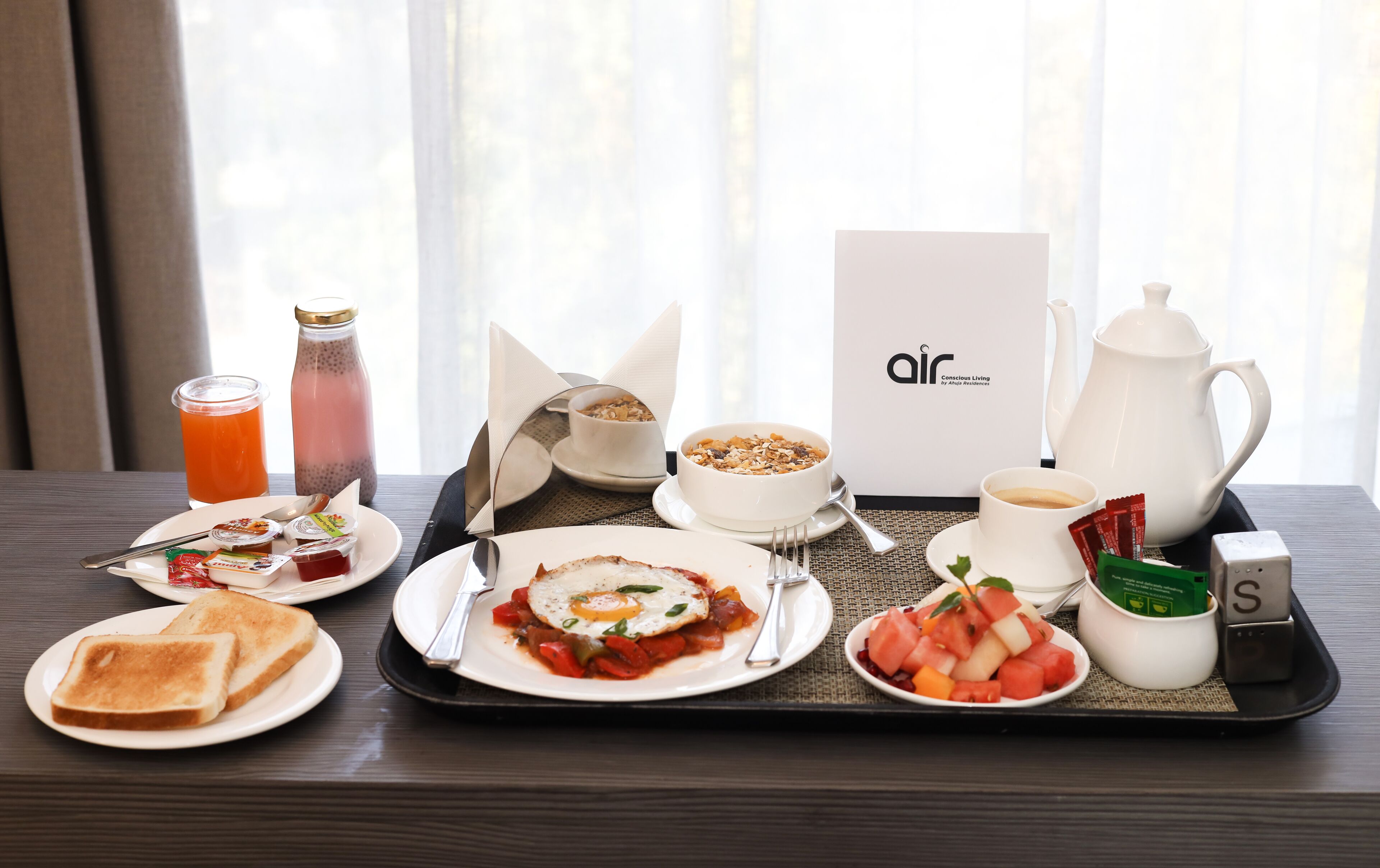 Una bandeja de desayuno con tostadas, huevos revueltos, un tazón de cereal, una taza de café, una botella de jugo y una tetera.